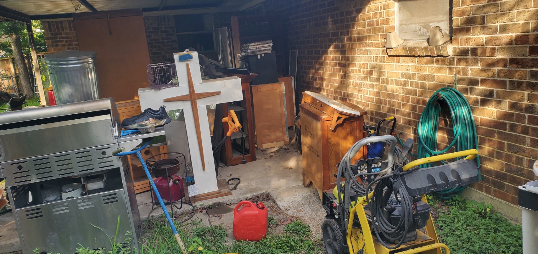 Before — Houston carport packed with fridge, cabinets, grill, cross, and garden hose before Holman Hauling cleanout